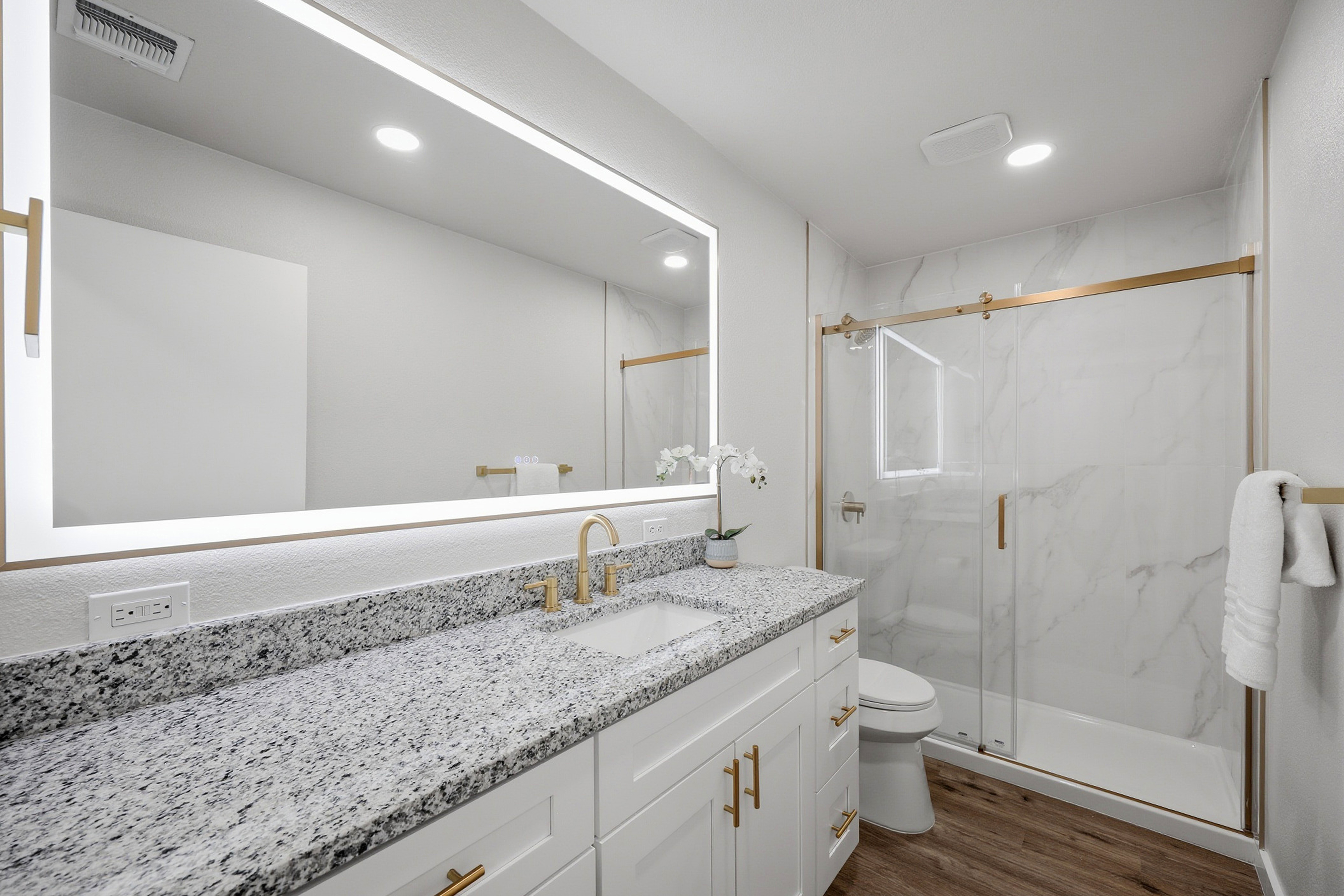 Master bathroom with LED backlit mirror, gold fixtures, granite double vanity, and glass-enclosed marble shower